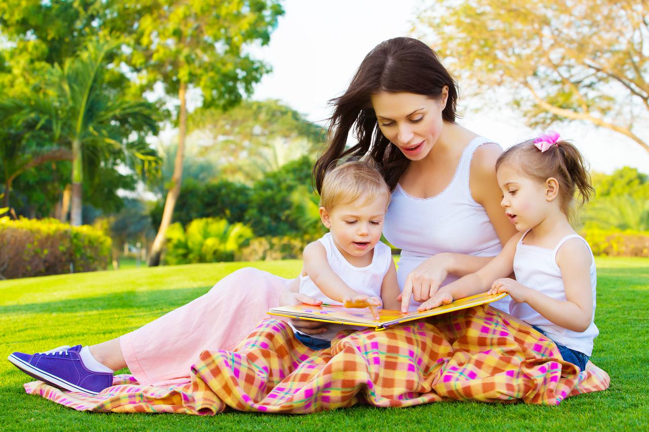 Photo of young brunette woman teaching two sweet kids, cheerful mother with cute daughter and lovely son read interesting fairytale, children enjoying book on backyard in daycare, spring season